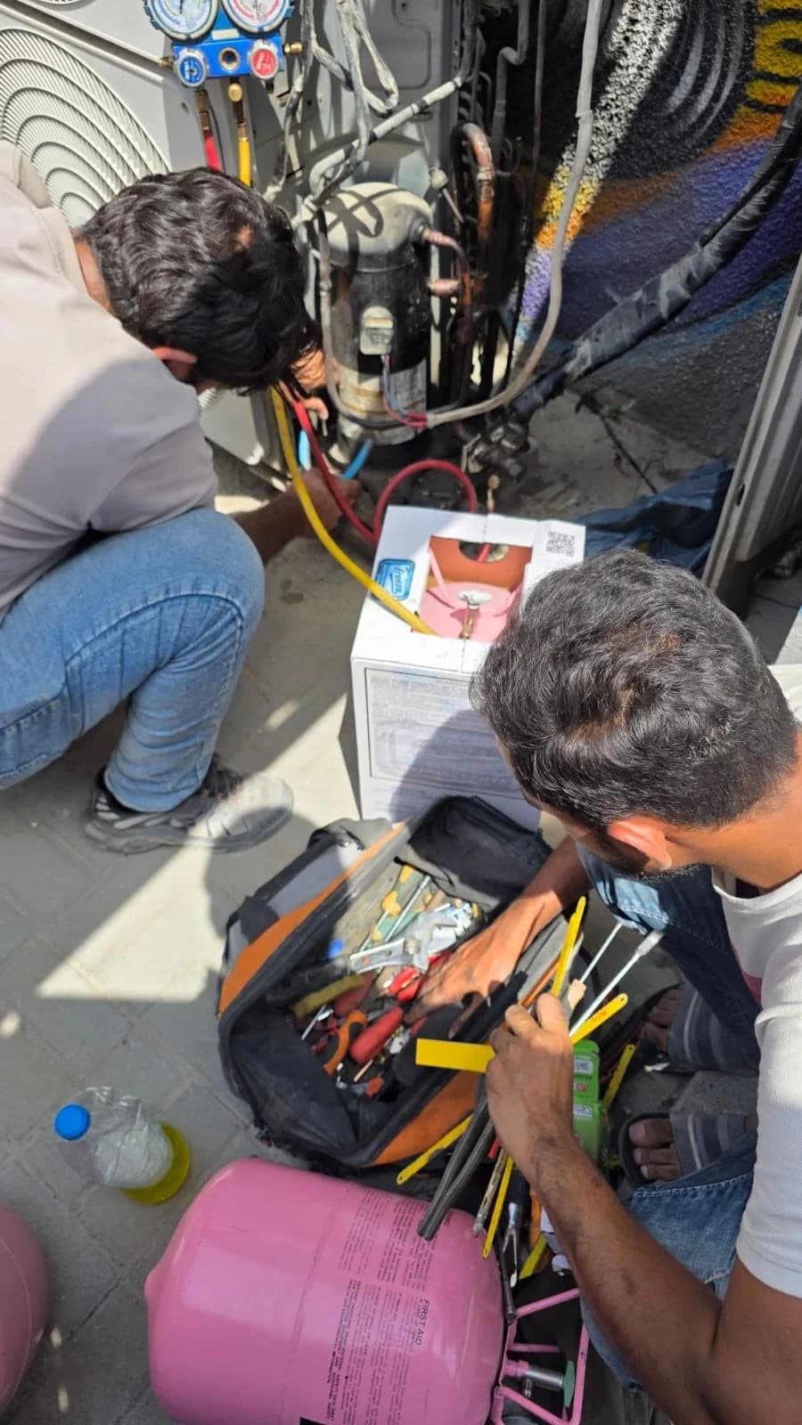 Technicians preparing wiring and refrigerant work beside an outdoor AC unit