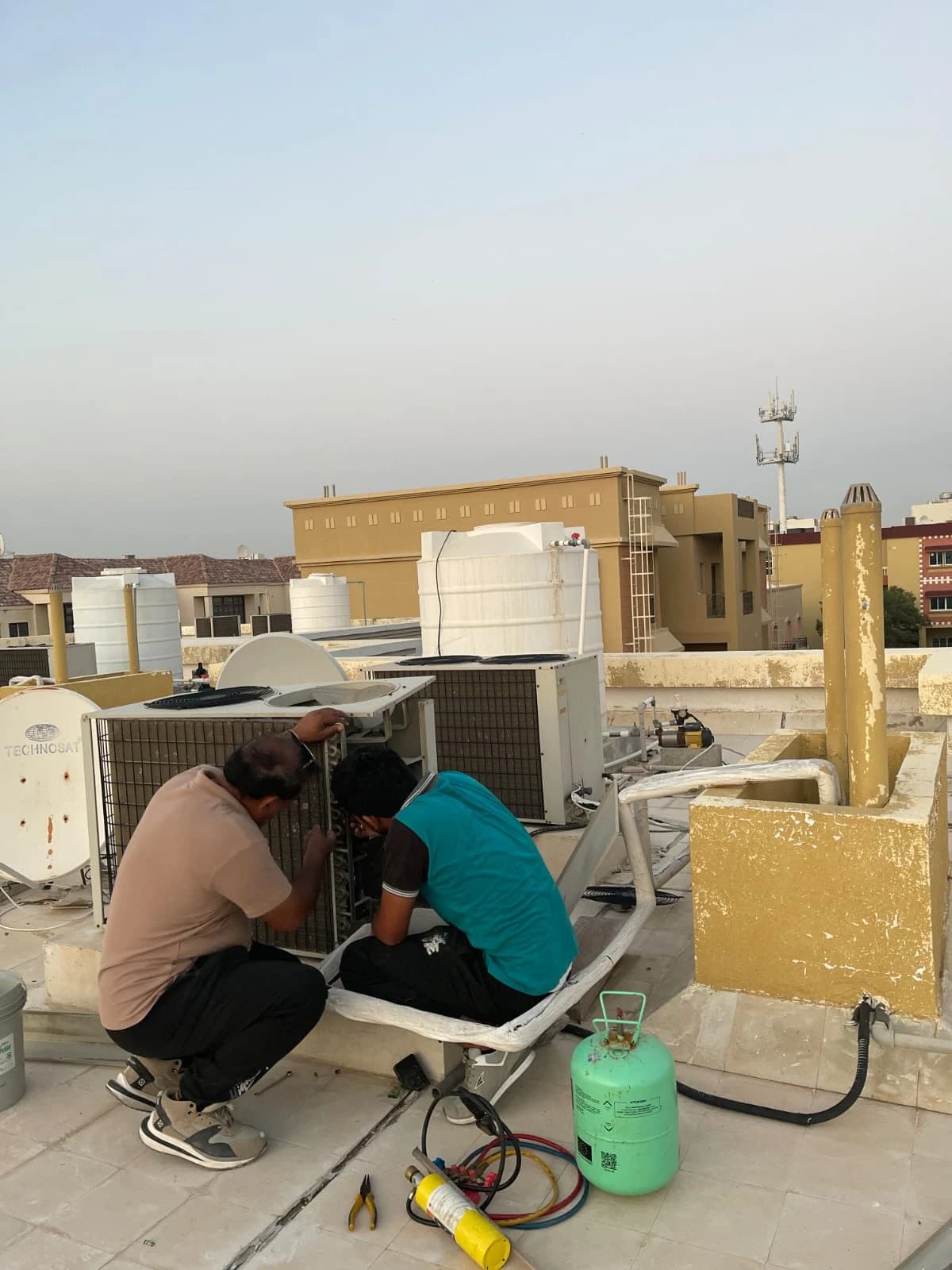 Technicians working at outdoor AC units on a rooftop with a refrigerant cylinder