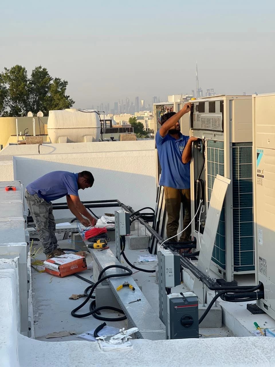 Two technicians installing rooftop AC units with the Dubai skyline in the background