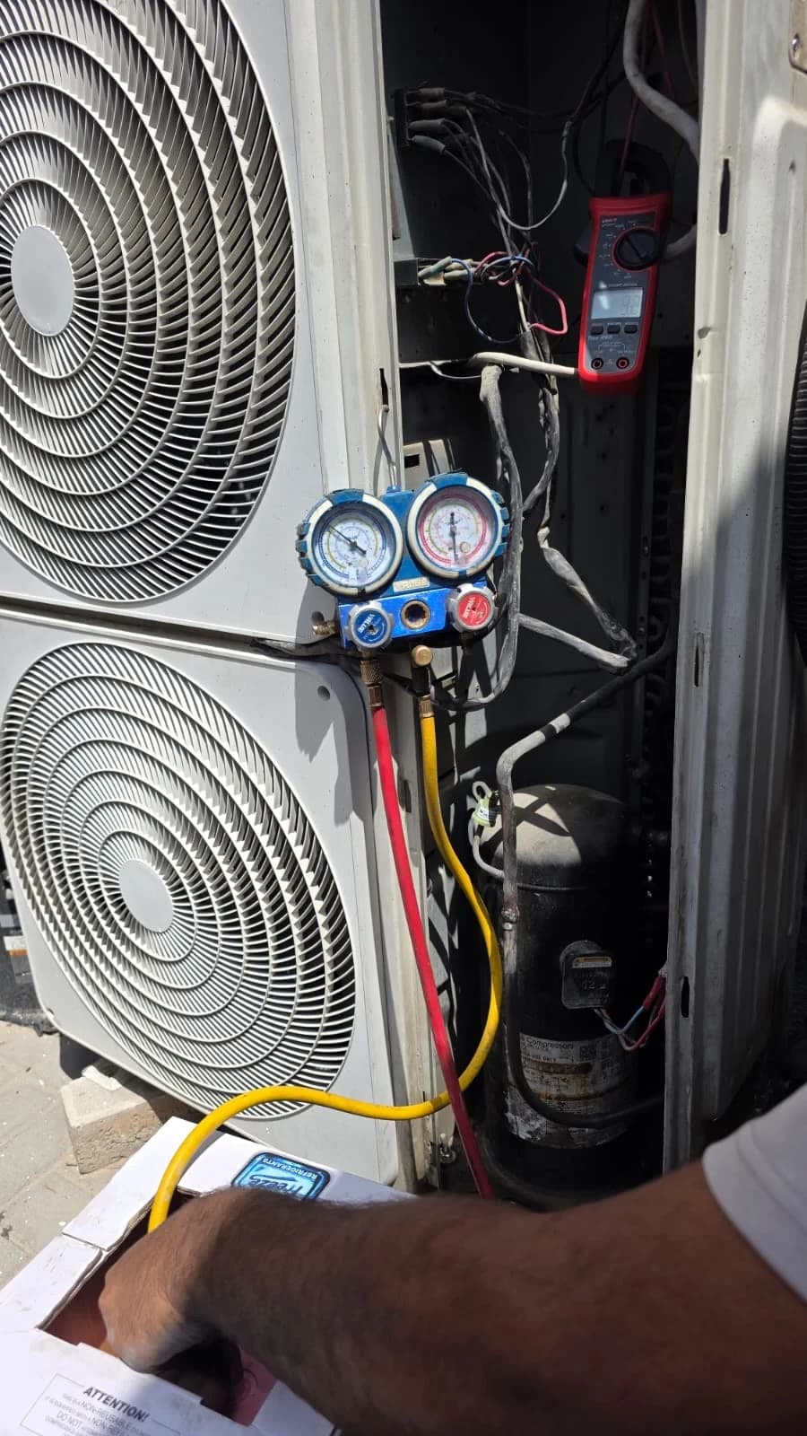 Open outdoor AC unit connected to pressure gauges and a multimeter during diagnosis