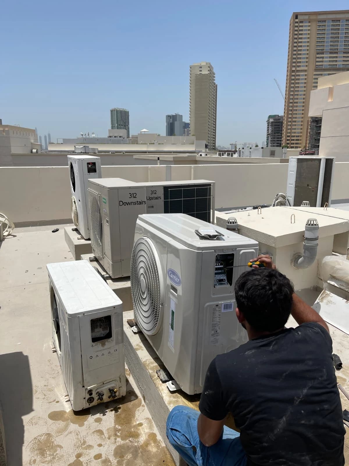 Technician servicing outdoor AC units on a rooftop with city towers behind